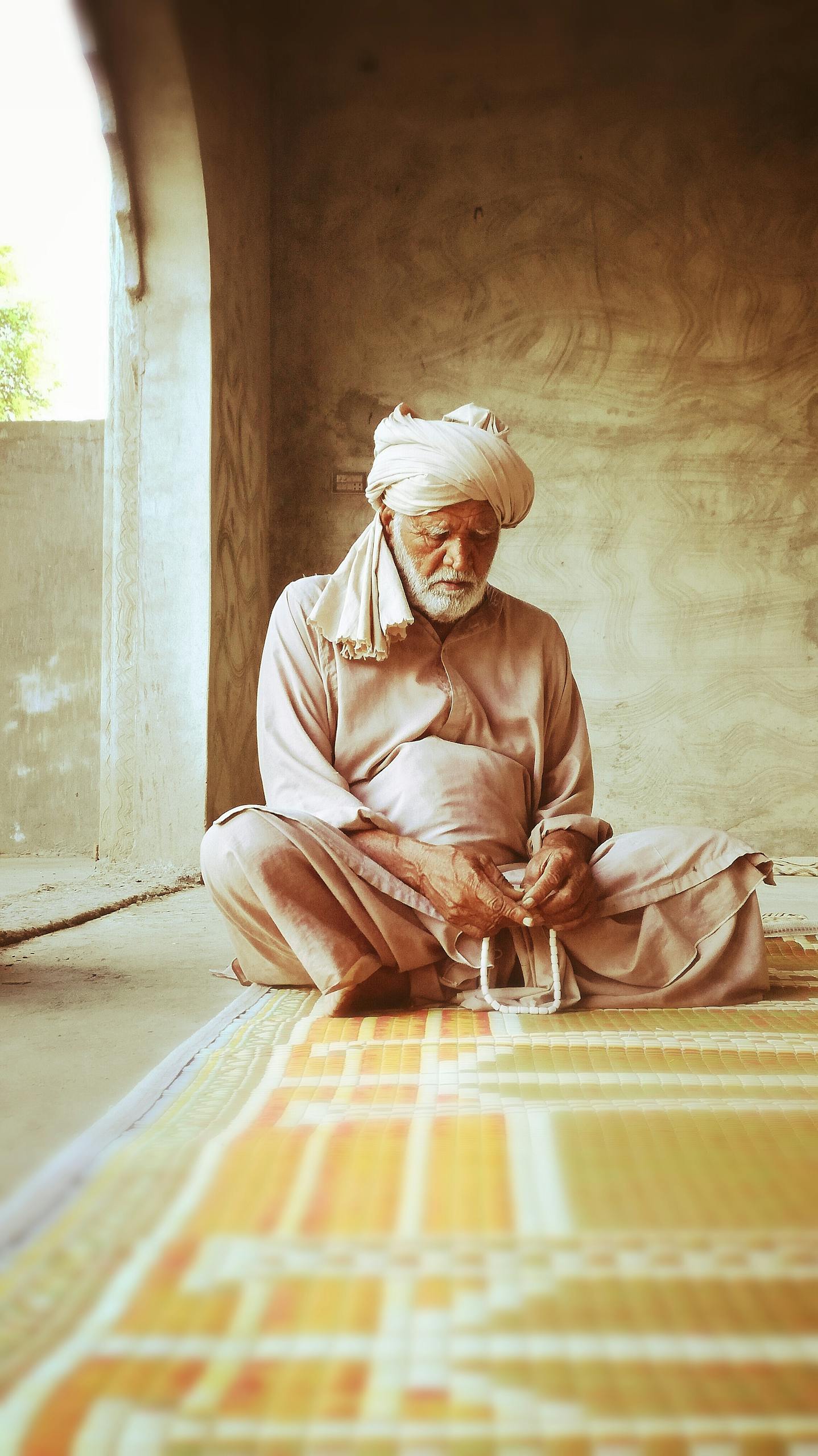Senior man in traditional attire sitting indoors on a colorful rug.
