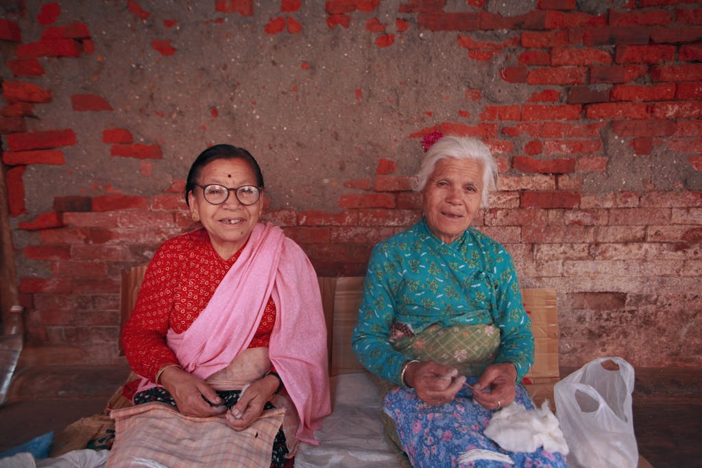 Two elderly women with vibrant attire sit smiling by a rustic brick wall.