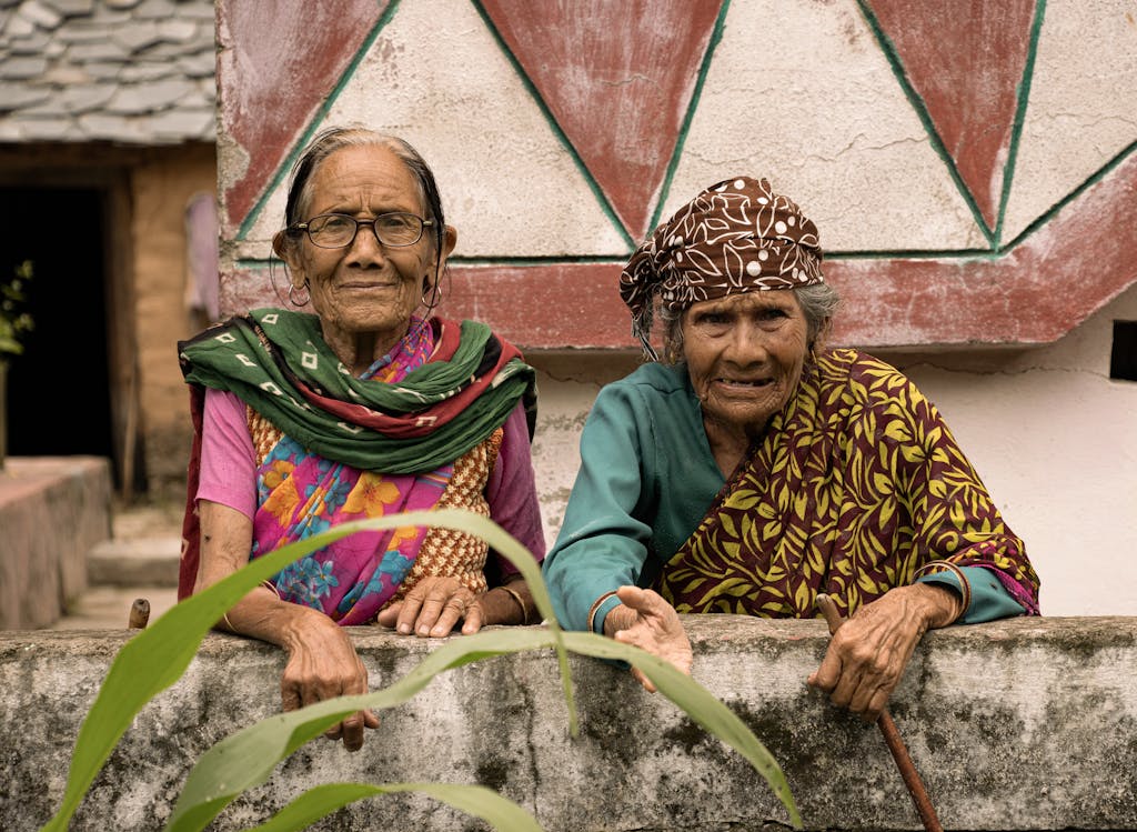 Two senior Indian women in colorful traditional attire leaning on a wall, smiling.